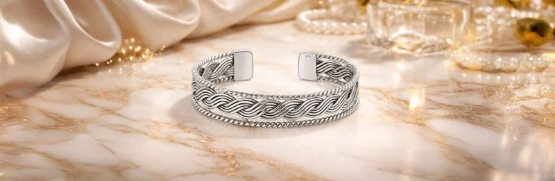 أسوارة فضة مفتوح بتصميم مجدول أنيق، معروض على خلفية فاخرة بدرجات الشمبانيا تضم قماش ساتان ناعم ولآلئ وزجاجة عطر، في مشهد يوحي بالفخامة والرقي.